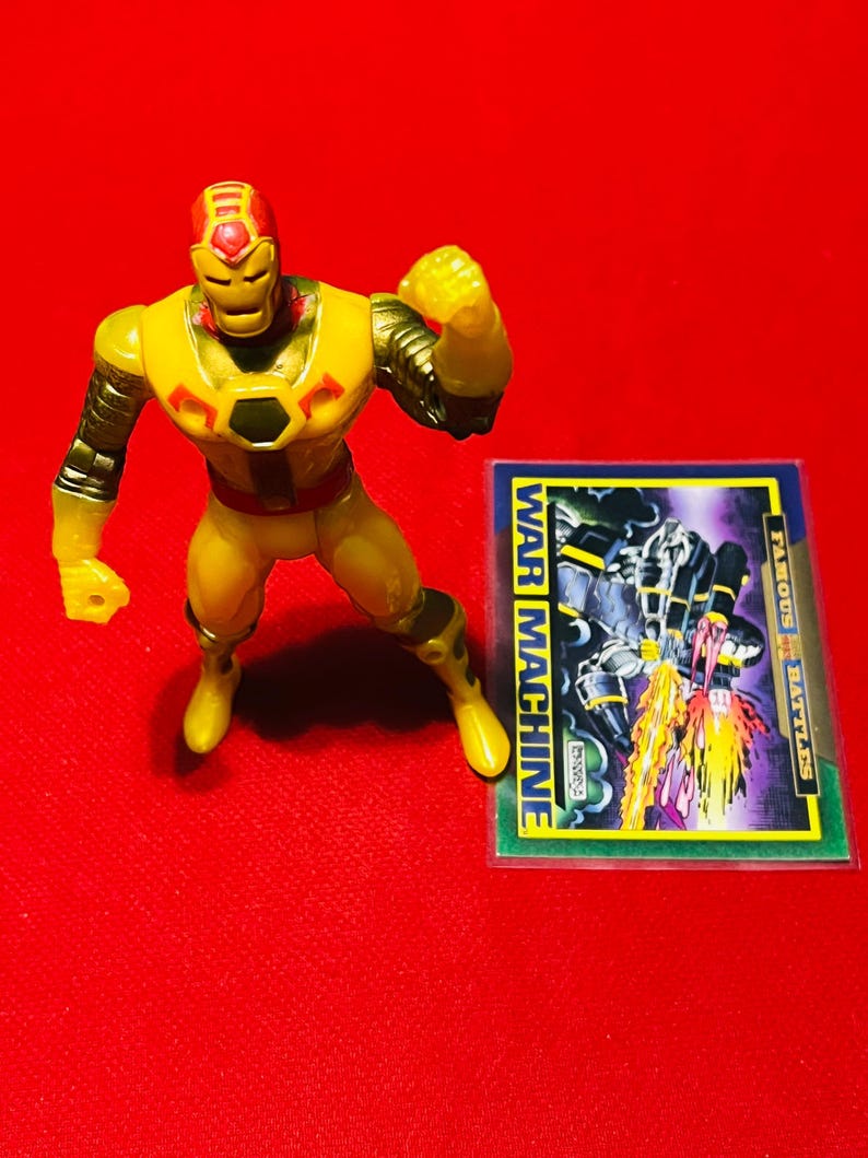 May include: Yellow and red plastic action figure of Iron Man's alter ego War Machine. The figure is posed with one arm raised and the other arm bent at the elbow. The figure is standing on a red surface. A trading card with the text "WAR MACHINE" is visible in the background.