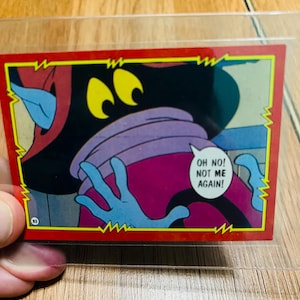 May include: A vintage trading card featuring a cartoon character with yellow eyes, a purple face, and blue hands. The character is saying "OH NO! NOT ME AGAIN!" in a speech bubble. The card has a red border.