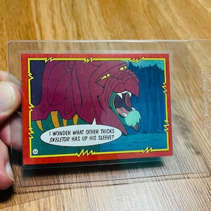 May include: A trading card featuring a red monster with a wide open mouth and sharp teeth. The card has a yellow border and the text "I WONDER WHAT OTHER TRICKS SKELETOR HAS UP HIS SLEEVE?" in a white speech bubble.