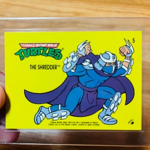 May include: A Teenage Mutant Ninja Turtles trading card featuring The Shredder. The card has a yellow background with a cartoon illustration of Shredder in blue and purple. The card also has the number 5 in the upper right corner.