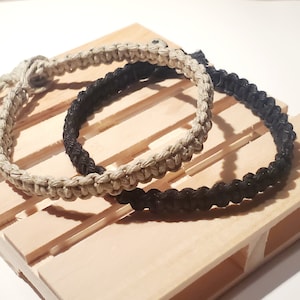 May include: Two macrame bracelets, one in natural beige and the other in black, rest on a small wooden pallet. The bracelets are made of woven cord, showcasing a textured, handcrafted appearance. The neutral tones of the bracelets contrast with the light wood.