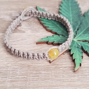 May include: A hemp bracelet with a yellow gemstone bead. The bracelet is on a wooden surface with a green leaf behind it.
