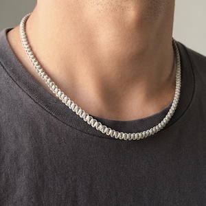 May include: A light gray, woven cord necklace featuring a continuous, repeating knot pattern. The necklace is constructed from a single strand of cord, showcasing a textured, braided design that sits closely around the neck area.