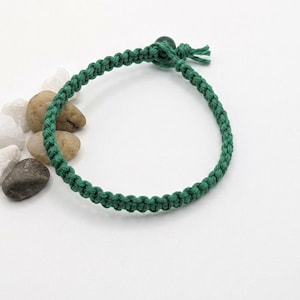 May include: A dark green cord bracelet featuring a braided macrame pattern. The bracelet is secured with a single dark green bead closure and a knotted cord end. The design consists of a continuous woven cord structure with a circular shape.