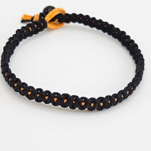 May include: A braided cord bracelet featuring a black exterior weave with a visible orange inner core. The bracelet includes a sliding knot closure mechanism finished with a single black bead and short orange cord ends.