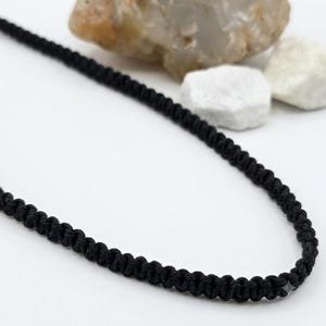 May include: A black cord necklace featuring a continuous series of intricate, hand-knotted macrame loops. The cord is constructed from a thin, dark fiber material, showcasing a uniform, repeating knot pattern throughout its entire length.
