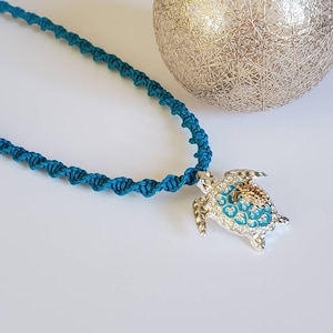 May include: Teal macrame necklace with a silver turtle pendant. The turtle pendant has blue and clear crystal accents. A silver decorative sphere is in the background.
