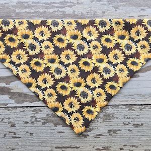 May include: A brown and black dog bandana with a sunflower pattern. The bandana is triangular in shape and has a yellow and black sunflower print.