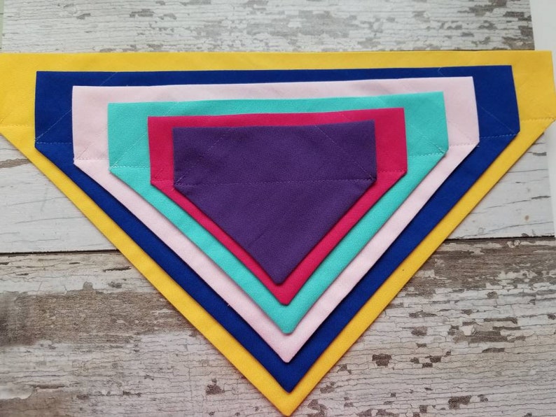 May include: A set of six fabric dog bandanas in various colors, including yellow, blue, pink, red, teal, and purple. The bandanas are layered on top of each other, with the largest bandana on the bottom and the smallest bandana on top.