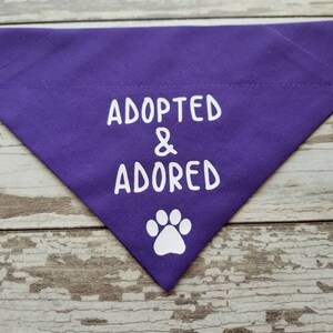 May include: Purple dog bandana with white text that reads "Adopted & Adored" and a white paw print.