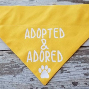 May include: Yellow dog bandana with white text that reads "Adopted & Adored" and a white paw print.
