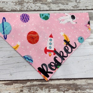 May include: Pink fabric dog bandana with a space theme. It features planets, stars, a rocket, and an astronaut. The bandana has the word "Rocket" written on it in black.