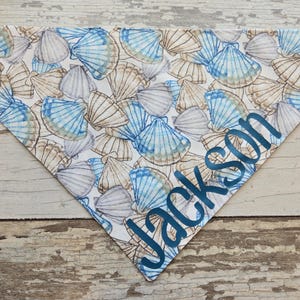 May include: A triangular pet bandana featuring a seashell pattern in shades of blue, tan, and white. The name "Jackson" is printed in blue script across the bottom corner. The bandana is designed to be worn around a pet's neck.