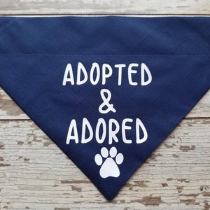 May include: A navy blue dog bandana with white text that reads "Adopted & Adored" and a white paw print.