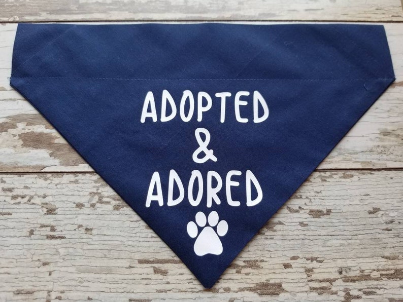 May include: A navy blue dog bandana with white text that reads "Adopted & Adored" and a white paw print.