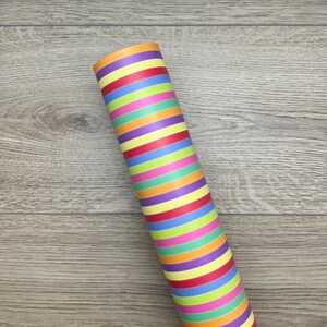 May include: A roll of colourful striped wrapping paper with red, orange, yellow, green, blue, purple and pink stripes. The paper is rolled up and lying on a wooden surface.