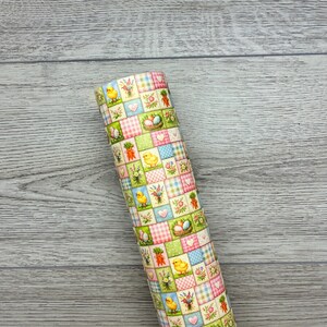 May include: Rolled fabric with a patchwork design featuring Easter illustrations. The pattern includes chicks, carrots, eggs in nests, and floral elements in a pastel colour palette of pink, green, and blue. The fabric is rolled up.