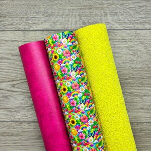 Peut inclure: Trois rouleaux de matériau de loisirs créatifs : un similicuir rose vif uni, un avec un motif vibrant de fleurs colorées, papillons, arcs-en-ciel et cerises, et une feuille à paillettes jaune vif avec une surface texturée et scintillante.