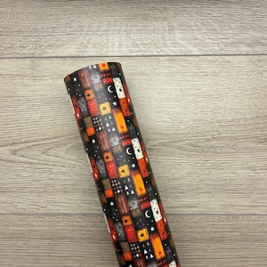 May include: A rolled-up sheet of patterned material featuring a patchwork design. The pattern includes squares in shades of red, orange, gray, and black, adorned with stars, moons, and geometric shapes. The material is displayed against a light wood grain background.