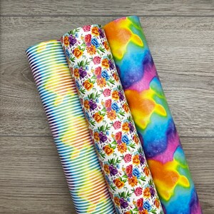 May include: Three rolls of colourful patterned fabric. The fabric on the left has rainbow stripes, the middle fabric has a floral pattern with orange, yellow, purple, and red flowers, and the fabric on the right has a tie-dye pattern with blue, orange, and pink colours.