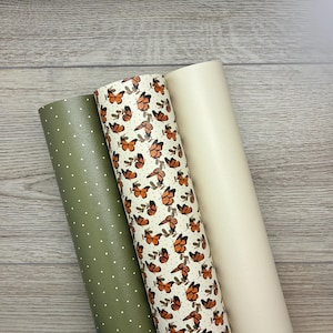 May include: Three rolls of faux leather fabric. One is olive green with white polka dots, the second is cream-colored, and the third is patterned with orange butterflies on a cream background. The rolls are arranged vertically.