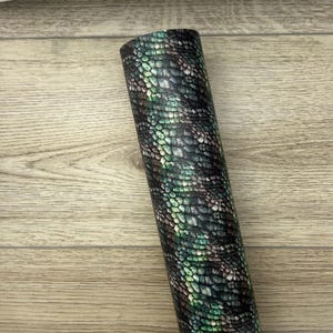 May include: A close-up of a green and black snakeskin patterned fabric. The fabric has a textured surface and is rolled up.