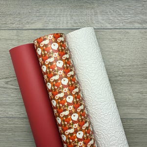 May include: Three rolls of decorative craft material. One is solid red, another features a pumpkin and floral pattern, and the third is white with a textured lace design. The rolls are displayed on a light gray wooden surface.