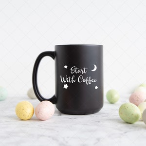 Pu&ograve; includere: Tazza da caff&egrave; nera con testo bianco che recita "Start With Coffee" e una mezzaluna e stelle. La tazza &egrave; su una superficie di marmo bianco con uova macchiate di colore pastello sparse intorno.