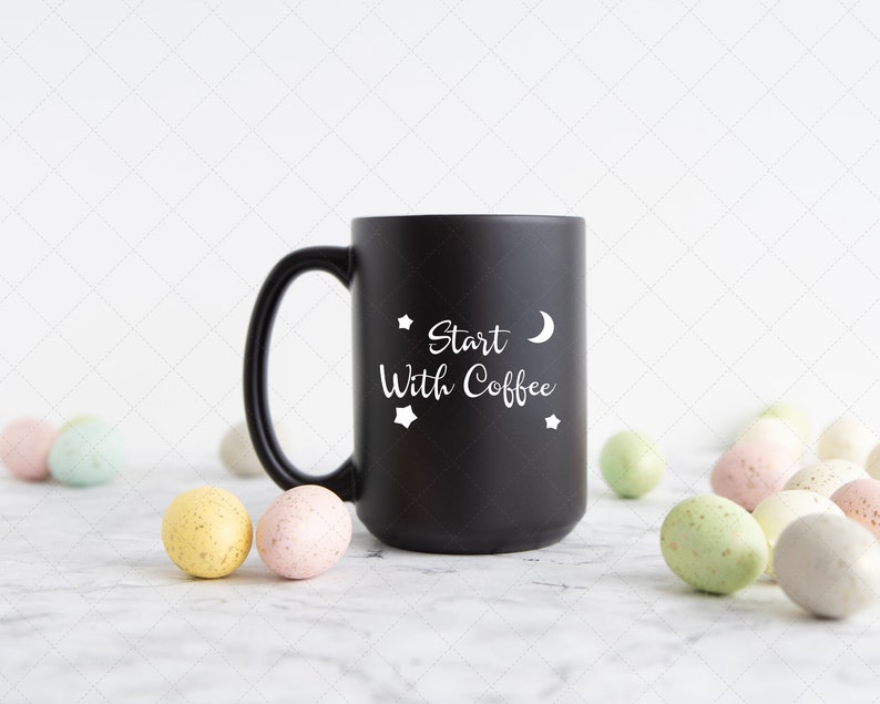 Pu&ograve; includere: Tazza da caff&egrave; nera con testo bianco che recita "Start With Coffee" e una mezzaluna e stelle. La tazza &egrave; su una superficie di marmo bianco con uova macchiate di colore pastello sparse intorno.