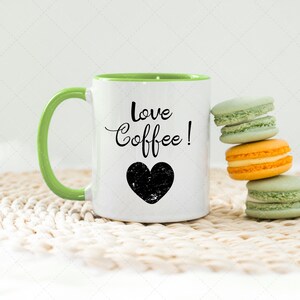 Pu&ograve; includere: Una tazza da caff&egrave; bianca con un bordo e un manico verdi. La tazza ha la scritta "love coffee!" e un cuore nero. La tazza &egrave; appoggiata su un sottopentola in crochet bianco accanto a una pila di quattro macarons verde pastello e arancioni.
