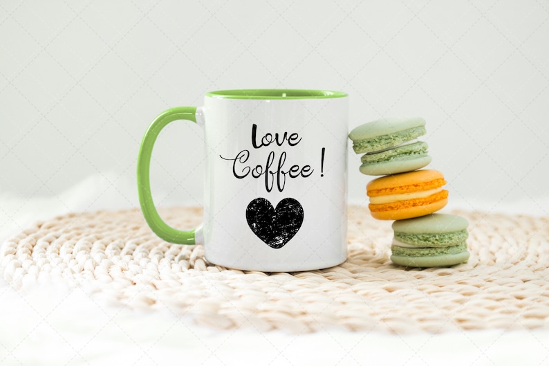 Pu&ograve; includere: Una tazza da caff&egrave; bianca con un bordo e un manico verdi. La tazza ha la scritta "love coffee!" e un cuore nero. La tazza &egrave; appoggiata su un sottopentola in crochet bianco accanto a una pila di quattro macarons verde pastello e arancioni.