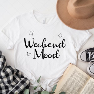 Pu&ograve; includere: Maglietta bianca con il testo "Weekend Mood" in corsivo nero. La maglietta &egrave; circondata da un cappello, un libro, una coperta a quadri e un rametto di verde.