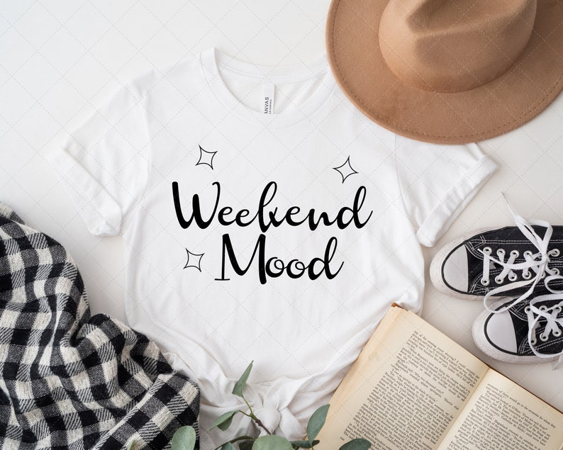 Pu&ograve; includere: Maglietta bianca con il testo "Weekend Mood" in corsivo nero. La maglietta &egrave; circondata da un cappello, un libro, una coperta a quadri e un rametto di verde.
