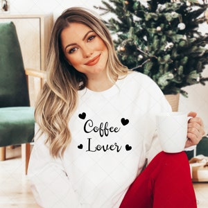 Pu&ograve; includere: Una felpa bianca con la scritta "Coffee Lover" in lettere nere e piccoli cuori neri. La modella indossa la felpa e tiene in mano una tazza bianca.