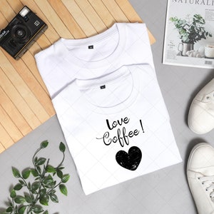 Pu&ograve; includere: Due magliette bianche, una con il testo "love coffee!" e una grafica a forma di cuore nero. Le magliette sono disposte su una superficie di legno con una macchina fotografica, piante e scarpe da ginnastica bianche.