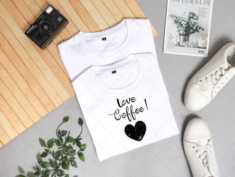 Pu&ograve; includere: Due magliette bianche, una con il testo "love coffee!" e una grafica a forma di cuore nero. Le magliette sono disposte su una superficie di legno con una macchina fotografica, piante e scarpe da ginnastica bianche.