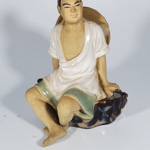 May include: A ceramic figurine of a man wearing a white robe and a straw hat, sitting on a rock. The figurine is painted in shades of brown, white, and green.