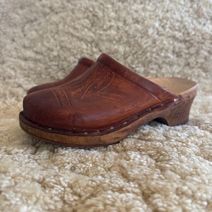 Könnte beinhalten: Ein Paar braune Lederclogs mit Holzsohlen. Die Clogs haben ein klassisches Design mit dekorativen Details auf der Vorderseite. Die Holzsohlen sind mit kleinen Holzstiften befestigt. Diese Clogs sind eine traditionelle Schuhart.