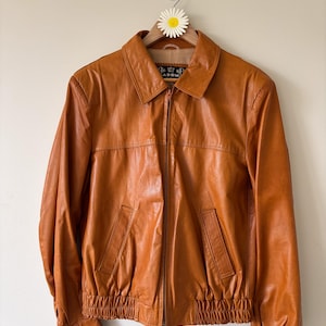 May include: A vintage, orange-brown leather jacket with a zip-up front, two front pockets, and a collared neckline. The jacket has a ribbed hem and cuffs, and is hanging on a wooden hanger with a daisy decoration.