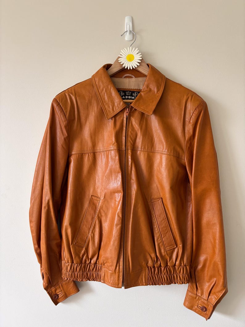 May include: A vintage, orange-brown leather jacket with a zip-up front, two front pockets, and a collared neckline. The jacket has a ribbed hem and cuffs, and is hanging on a wooden hanger with a daisy decoration.