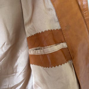 May include: Close-up of the interior of a brown leather jacket. The jacket features a tan lining and a small pocket with a decorative, scalloped edge. The leather has a slight sheen, and the zipper is visible on the right side.
