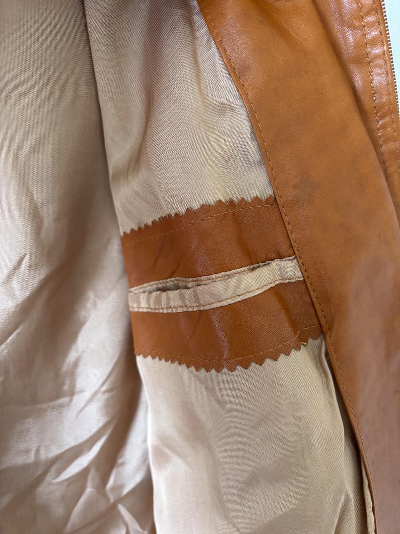 May include: Close-up of the interior of a brown leather jacket. The jacket features a tan lining and a small pocket with a decorative, scalloped edge. The leather has a slight sheen, and the zipper is visible on the right side.