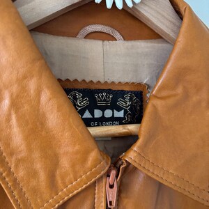 May include: Close-up of a tan leather jacket with a zipper closure. The jacket has a wide collar and a label that reads "ADOM OF LONDON". The jacket is hanging on a wooden hanger with a daisy decoration.