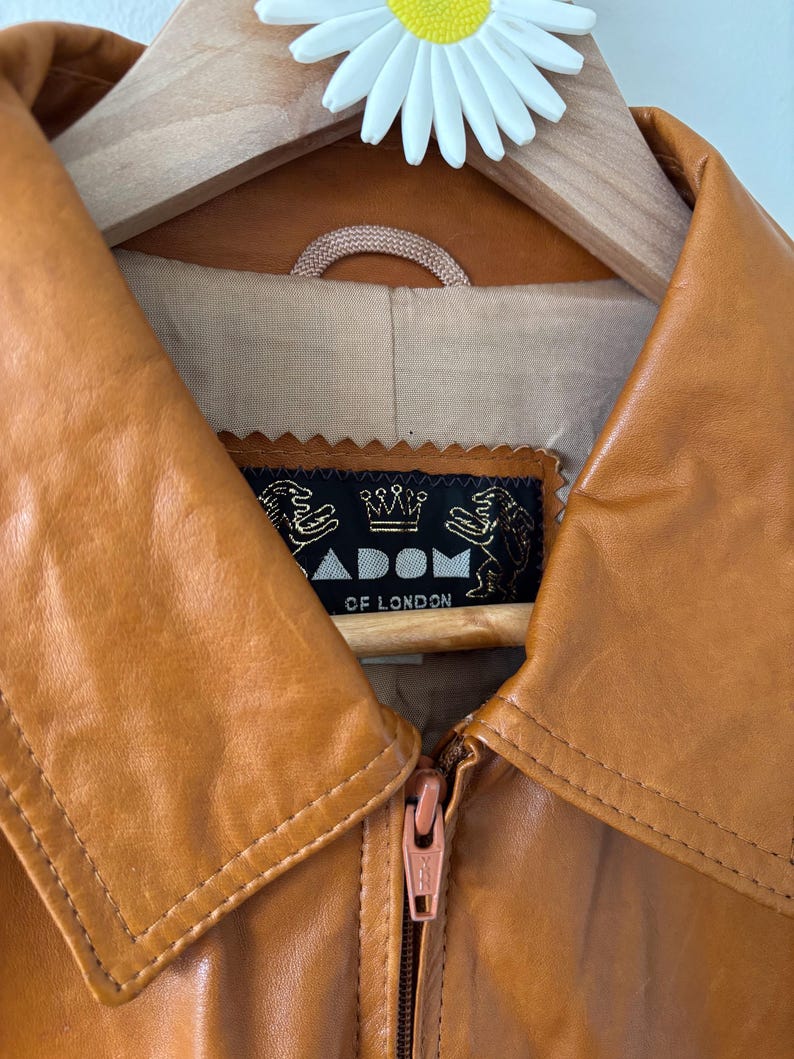 May include: Close-up of a tan leather jacket with a zipper closure. The jacket has a wide collar and a label that reads "ADOM OF LONDON". The jacket is hanging on a wooden hanger with a daisy decoration.