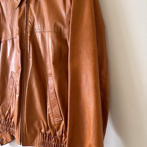 May include: A vintage brown leather jacket with a collared neckline and a zipper closure. The jacket features two front pockets and elastic cuffs and waistband. The leather has a glossy finish and a classic, timeless style.