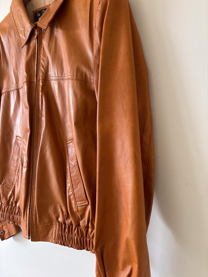 May include: A vintage brown leather jacket with a collared neckline and a zipper closure. The jacket features two front pockets and elastic cuffs and waistband. The leather has a glossy finish and a classic, timeless style.