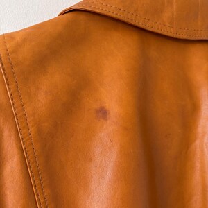 May include: Close-up of a tan leather jacket, showing the collar and back. The jacket has visible stitching and a small stain on the back. The leather has a smooth texture and a warm, earthy tone.