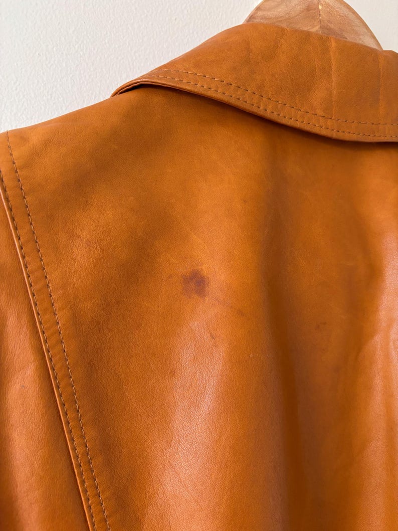 May include: Close-up of a tan leather jacket, showing the collar and back. The jacket has visible stitching and a small stain on the back. The leather has a smooth texture and a warm, earthy tone.