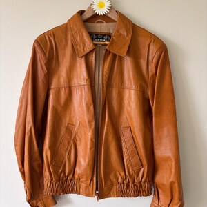 May include: A tan leather jacket with a collared neckline, front zipper, and two front pockets. The jacket has a ribbed hem and cuffs, and is hanging on a wooden hanger with a daisy decoration.