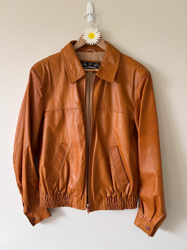 May include: A tan leather jacket with a collared neckline, front zipper, and two front pockets. The jacket has a ribbed hem and cuffs, and is hanging on a wooden hanger with a daisy decoration.